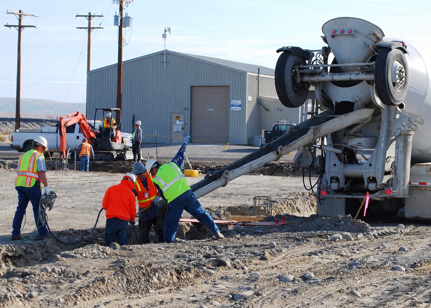 Workers pouring concrete footings beside a mixer truck and compact excavator at Hanford, Washington